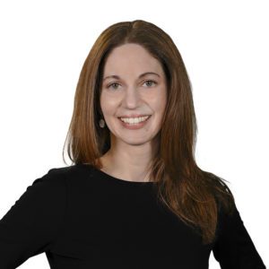 A person with long brown hair is smiling and wearing a black top against a white background.