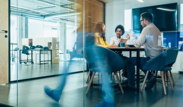 Businesspeople in Conference Room and Colleagues Walking By. Blurred motion of colleagues walking briskly down office hallway as colleagues sit in conference room discussing ideas. Busy modern workplace. Working process in the office, business people working, walking and talking, blurred motion