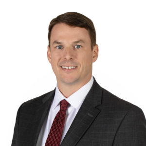 A person in a dark suit with a white shirt and red tie is smiling against a white background.