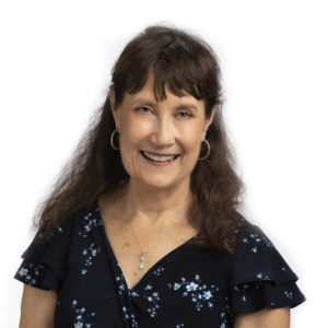 A woman with long brown hair is smiling and wearing a black floral dress with hoop earrings against a white background.