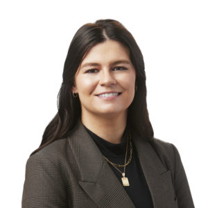 A person wearing a brown blazer and gold necklaces smiles against a white background.