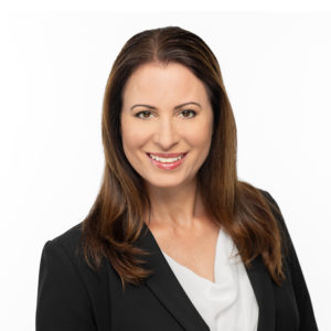 A woman with long brown hair is smiling and wearing a black blazer over a white top against a plain white background.