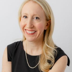 A woman with long blonde hair and a black sleeveless top is smiling and wearing a pearl necklace.