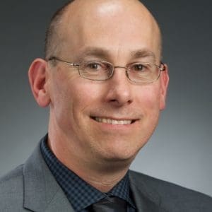 A person is wearing glasses, a suit, and a tie, and is smiling against a plain dark background.