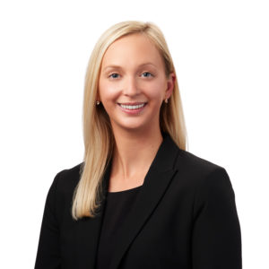 A woman with long, straight blonde hair in a professional black blazer smiles against a plain white background.