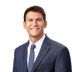 A person wearing a dark suit, white shirt, and blue tie is smiling against a white background.