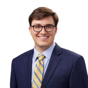 A person is wearing a dark blue suit, light blue dress shirt, and a yellow-striped tie, smiling at the camera against a white background.