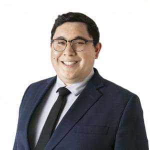 A person wearing glasses and a blue suit with a black tie smiles against a plain white background.