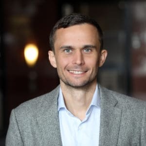 A man smiles while wearing a light blue shirt and a gray blazer.