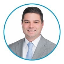 A person is smiling, dressed in a gray suit, white shirt, and light blue tie, inside a blue circular frame on a plain white background.
