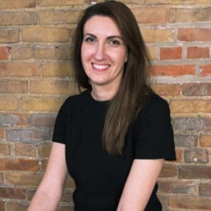 A person with long brown hair is smiling and standing in front of a brick wall.