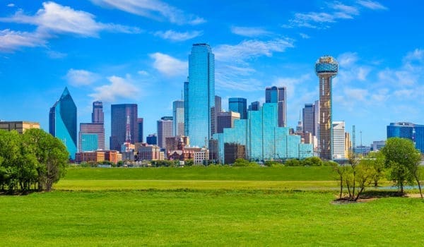 A city skyline featuring modern skyscrapers and a distinctive observation tower is set against a bright blue sky with a foreground of grassy parkland.