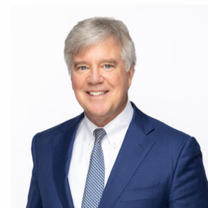 A man with gray hair wearing a blue suit, white shirt, and patterned tie is smiling.