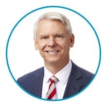 A smiling person with short gray hair is wearing a suit and striped tie, framed within a blue circle.