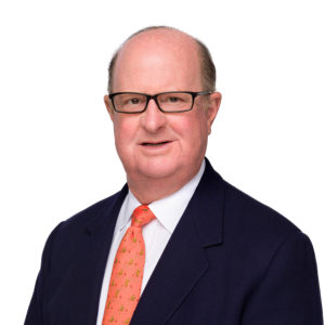 A person wearing glasses and a suit with a patterned orange tie stands against a plain white background.