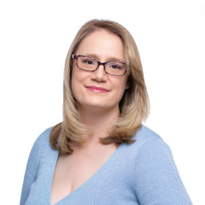 A person with blonde hair and glasses is smiling, wearing a light blue top against a white background.