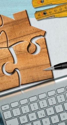 Wooden jigsaw puzzle pieces forming a house shape, on a desk with blank sheet of paper with copy space, calculator, folding ruler, drawing compass, Euro coins, pencil and a computer keyboard.