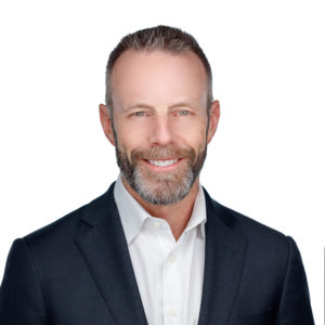 A man with a beard is wearing a white shirt and dark blazer, smiling against a plain white background.