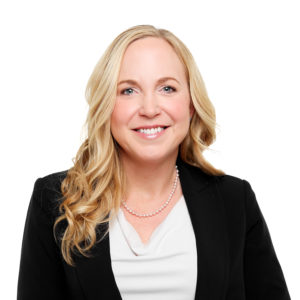 A woman with blonde hair is smiling and wearing a black blazer over a white top, accessorized with a pearl necklace, against a white background.