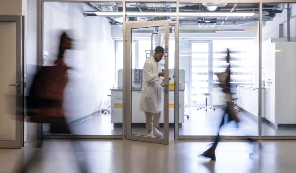 A chemical worker in a white coat stands between the doors of the faculty laboratory, students rush past him in the corridor. Blurred people while moving on the photo.