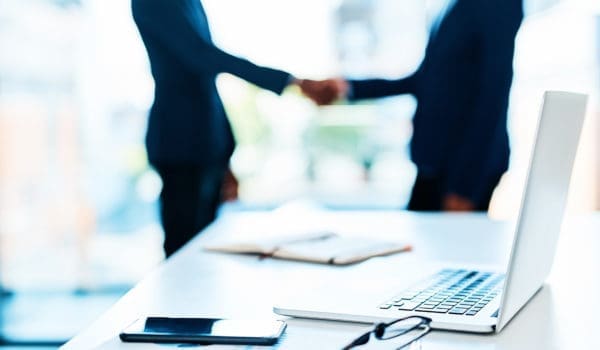 Cropped shot of a laptop on a desk with two businesspeople shaking hands in the background