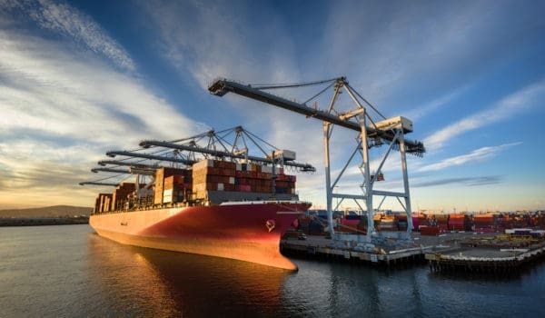 Aerial shot of a massive cargo ship loaded with containers docked in the Port of Long Beach, with the booms of ship-to-shore gantry cranes reaching out from the pier of the terminal.  

Authorization was obtained from the Port of Long Beach Security Division and POLB Harbor Patrol for this operation in restricted airspace.