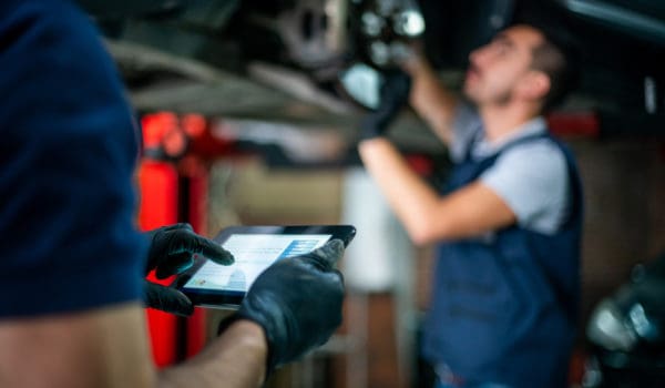 Focus on foreground of engineer holding a tablet checking the readings of the carâs sensor while the mechanic adjusts them on the disk - Car industry concepts