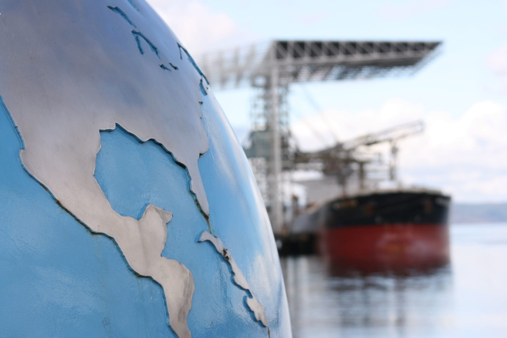 A globe with focus on the United States (USA) and Central America contrsted against a blurred background of a bulk cargo ship exemplifying the global nature of the cargo industry. Look at more